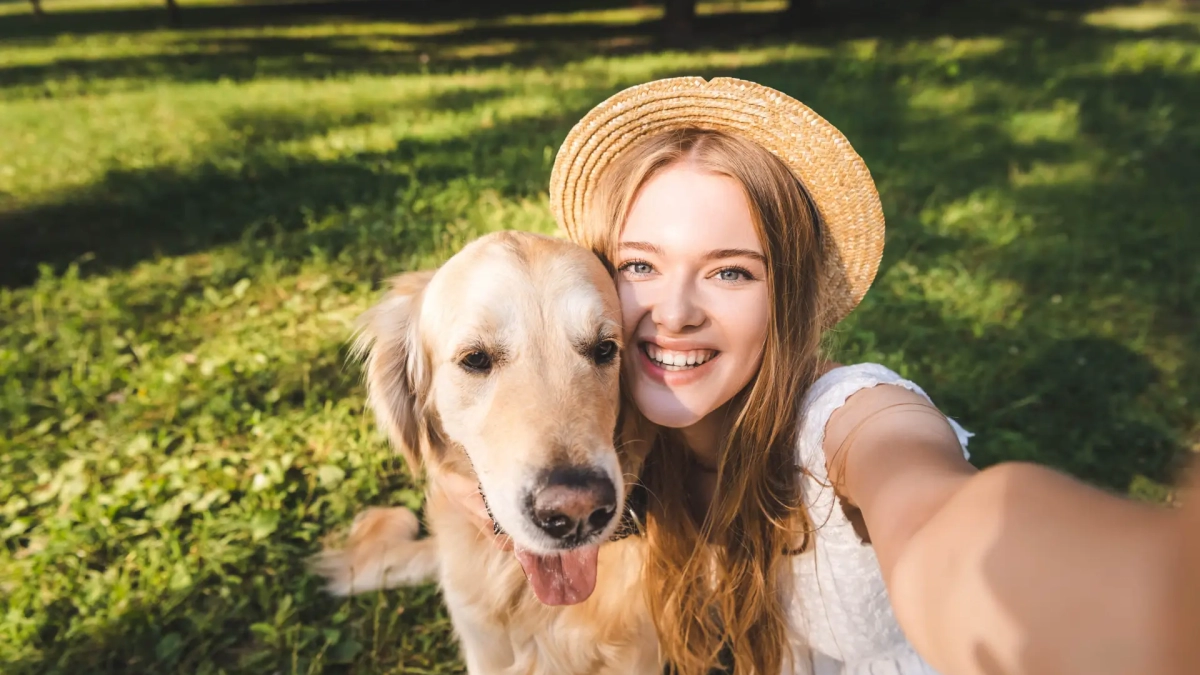 beautiful young girl in white dress and straw hat hugging golden retriever and taking selfie while sitting on meadow, smiling and looking at camera